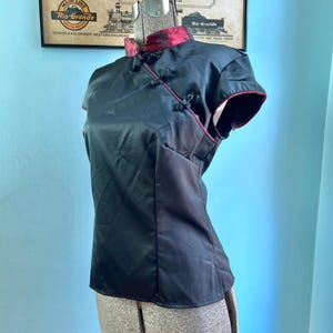May include: A black short-sleeved blouse featuring a mandarin collar with a red floral pattern. The garment includes a side-fastening design with three decorative knot buttons and red piping along the collar, sleeve edges, and front closure.