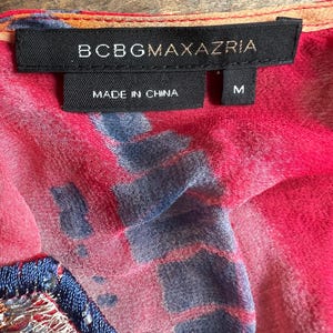 Peut inclure: Gros plan sur une &eacute;tiquette de v&ecirc;tement avec la marque BCBGMAXAZRIA en texte blanc sur une &eacute;tiquette noire. L'&eacute;tiquette indique &eacute;galement "MADE IN CHINA" et porte la taille "M". Le tissu est rose vif avec des motifs bleus et orange.