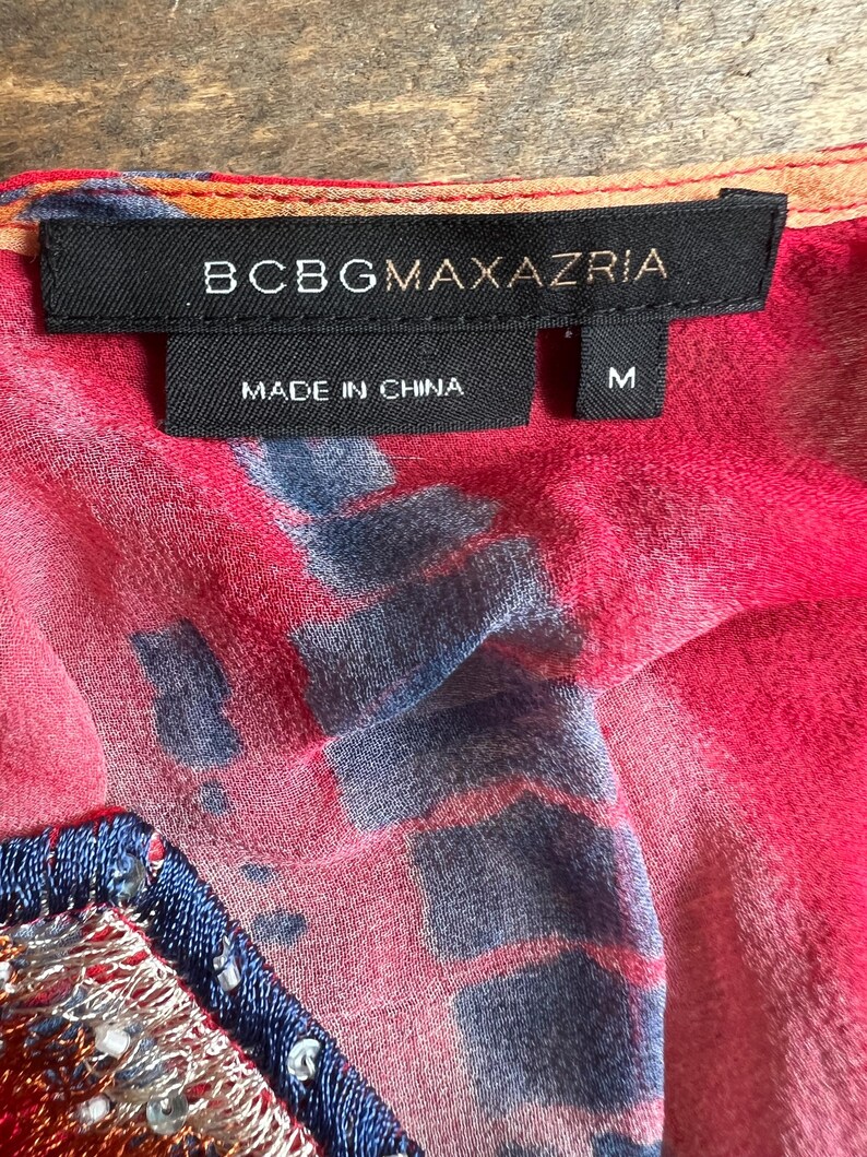Peut inclure: Gros plan sur une &eacute;tiquette de v&ecirc;tement avec la marque BCBGMAXAZRIA en texte blanc sur une &eacute;tiquette noire. L'&eacute;tiquette indique &eacute;galement "MADE IN CHINA" et porte la taille "M". Le tissu est rose vif avec des motifs bleus et orange.