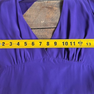 May include: A close-up of a vibrant purple dress with a V-neck design. The dress is measured with a yellow tape measure, showing a width of approximately 15 inches. The fabric appears to be a lightweight material.