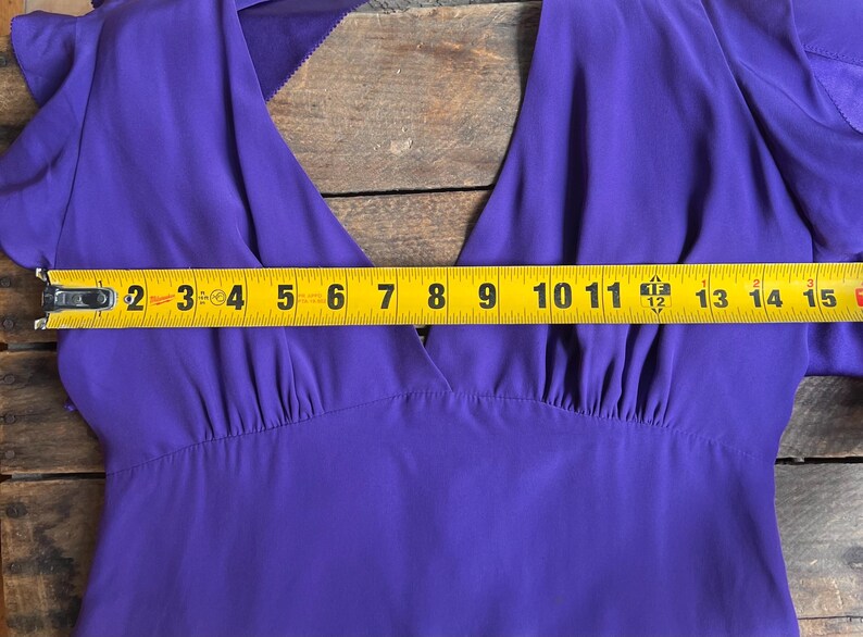 May include: A close-up of a vibrant purple dress with a V-neck design. The dress is measured with a yellow tape measure, showing a width of approximately 15 inches. The fabric appears to be a lightweight material.