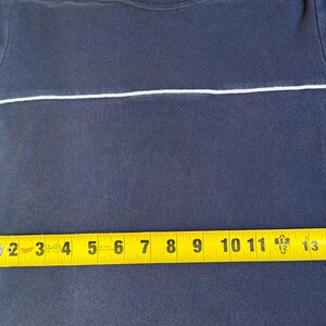 K&ouml;nnte beinhalten: Marineblaues T-Shirt mit einem wei&szlig;en horizontalen Streifen &uuml;ber der Brust. Ein gelbes Ma&szlig;band zeigt, dass das Shirt etwa 40 cm lang ist. Das Shirt hat kurze &Auml;rmel mit blauen und wei&szlig;en Akzenten.