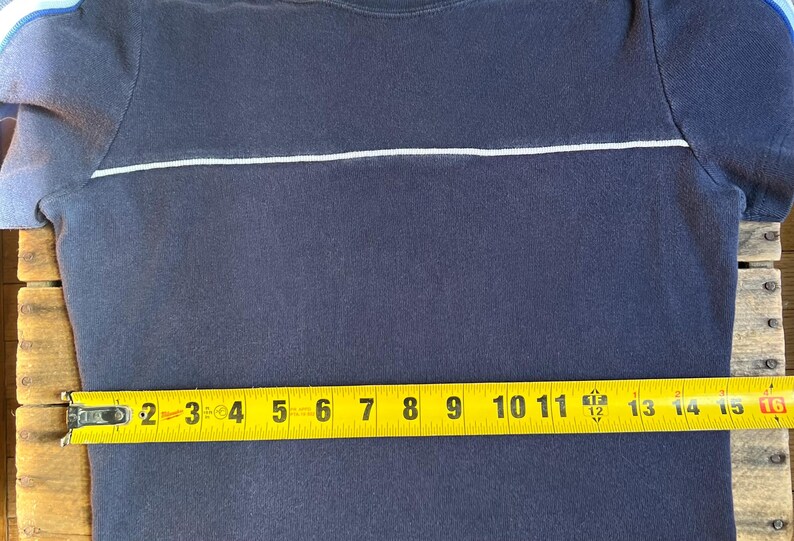 K&ouml;nnte beinhalten: Marineblaues T-Shirt mit einem wei&szlig;en horizontalen Streifen &uuml;ber der Brust. Ein gelbes Ma&szlig;band zeigt, dass das Shirt etwa 40 cm lang ist. Das Shirt hat kurze &Auml;rmel mit blauen und wei&szlig;en Akzenten.