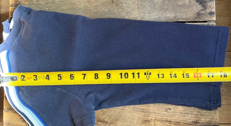 K&ouml;nnte beinhalten: Ein marineblaues Langarmshirt mit hellblau-wei&szlig;em Streifen am &Auml;rmel. Das Shirt wird mit einem gelben Ma&szlig;band gemessen und hat eine L&auml;nge von ca. 40 cm.