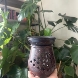 May include: A dark brown stone oil burner with a smooth, wide top basin and a rounded body featuring numerous circular cutouts throughout the surface, designed to allow light to pass through from a candle placed inside the central chamber.