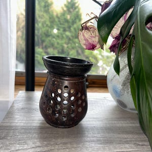 May include: A dark brown essential oil diffuser with a black top. The diffuser has a series of circular holes and sits on a gray surface. A vase with flowers and a green plant are in the background.