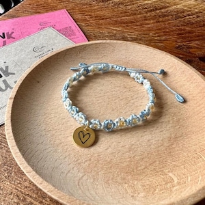 May include: A macrame bracelet with blue and white floral pattern and a gold-colored charm with a heart design. The bracelet is on a wooden plate, with pink and gray cards in the background.
