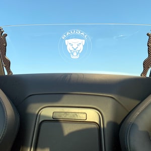 Puede incluir: Un interior de cuero negro de un coche con un deflector de viento transparente. El deflector tiene un logotipo de Jaguar blanco con el texto "JAGUAR" encima. El deflector está sujeto al coche con dos soportes de fibra de carbono negros.