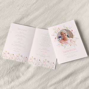 Puede incluir: Un programa de servicio conmemorativo de color rosa claro, abierto para mostrar el orden del servicio y una fotografía de una mujer sonriente. El programa está decorado con un borde floral y una corona floral alrededor de la foto.