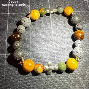 May include: A bracelet with brown, yellow, and green beads, interspersed with black lava rock beads. A silver cross charm is attached to the bracelet.