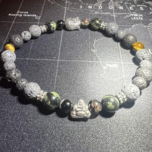 May include: A beaded bracelet with black lava rock beads, green and black gemstone beads, and two silver Buddha charms.