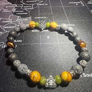 May include: A beaded bracelet featuring black lava rock, tiger's eye, and green jade beads. A silver charm of a meditating Buddha is positioned in the centre of the bracelet.