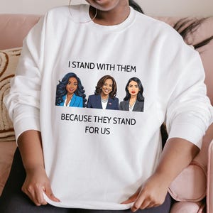 Op de afbeelding: Witte sweatshirt met een afbeelding van drie vrouwen en de tekst "I STAND WITH THEM BECAUSE THEY STAND FOR US". De vrouwen in cartoonstijl dragen businesspakken. Deze casual sweatshirt is geschikt voor dagelijks gebruik en is een comfortabel kledingstuk.