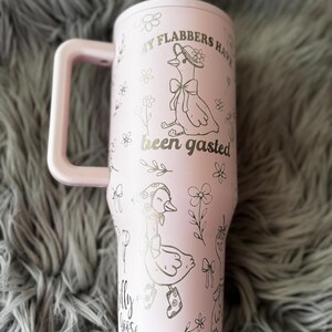 May include: A light pink tumbler with a handle, featuring illustrations of geese and floral designs. The text "My Flabbers Have Been Gasted" and "Silly Goose" are printed on the tumbler. The tumbler is on a gray, fuzzy surface.