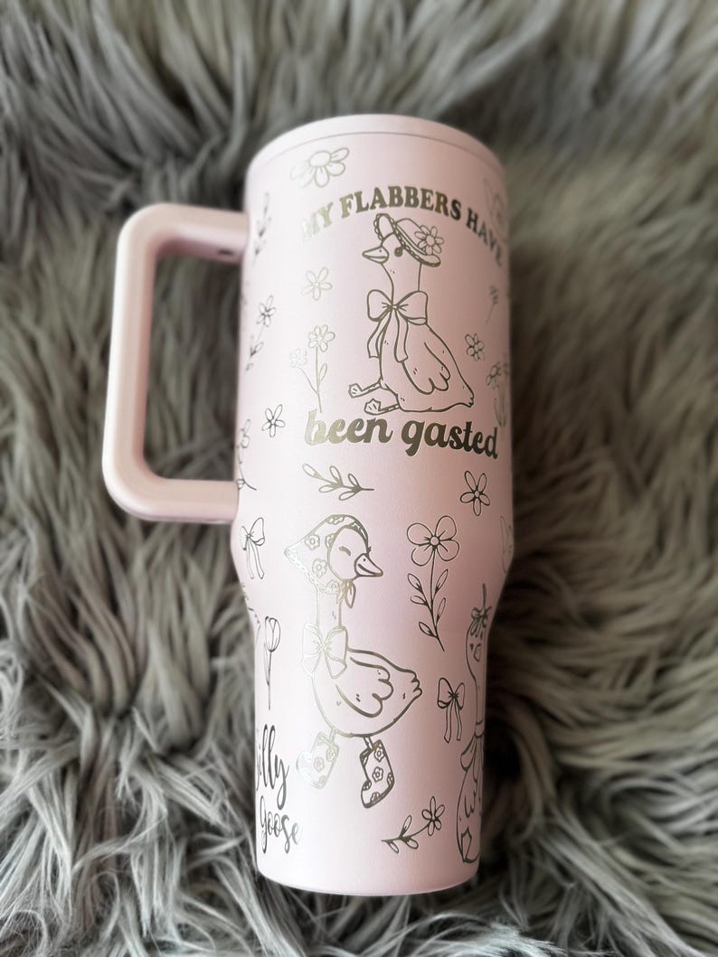 May include: A light pink tumbler with a handle, featuring illustrations of geese and floral designs. The text "My Flabbers Have Been Gasted" and "Silly Goose" are printed on the tumbler. The tumbler is on a gray, fuzzy surface.