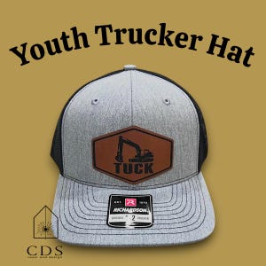 Pode incluir: Boné de caminhoneiro juvenil cinza e preto com um patch de couro marrom com um gráfico de escavadeira e o nome "TUCK". O boné tem uma frente estruturada e uma parte traseira de malha. O texto "Youth Trucker Hat" está arqueado acima.