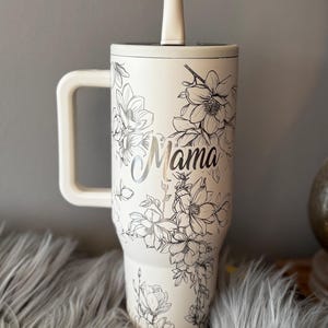 May include: Cream-colored tumbler with a handle and a straw, featuring a floral design and the word "Mama" in silver script. The tumbler has a white handle and straw, and the floral design is in black. The tumbler is designed to hold a large quantity of liquid.
