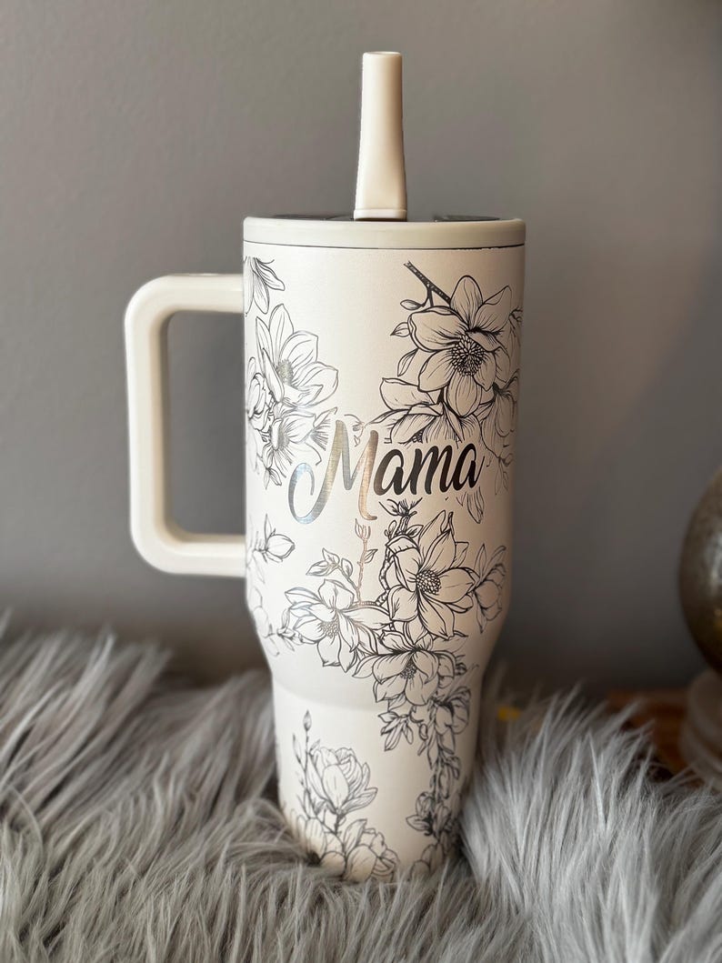 May include: Cream-colored tumbler with a handle and a straw, featuring a floral design and the word "Mama" in silver script. The tumbler has a white handle and straw, and the floral design is in black. The tumbler is designed to hold a large quantity of liquid.