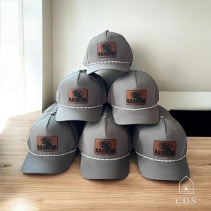 May include: A stack of gray baseball caps with a brown leather patch featuring the text "WIES" and a graphic. Each cap has a white and gray rope detail around the brim. The caps are stacked on a wooden surface.