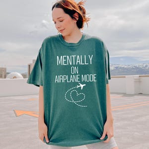 May include: A teal green t-shirt with the words "MENTALLY ON AIRPLANE MODE" in white, and a white airplane and heart graphic. The shirt has a relaxed fit and is made of a soft material.