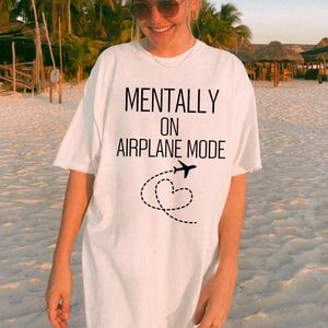 May include: White t-shirt featuring the text "MENTALLY ON AIRPLANE MODE" in black. A black airplane graphic with a dotted heart-shaped trail is below the text. The shirt has a loose fit.