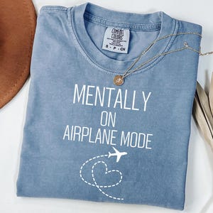 May include: A pale blue t-shirt with the text "MENTALLY ON AIRPLANE MODE" in white. A small airplane graphic and a dotted heart shape are below the text. A gold necklace with a pendant rests on the shirt.