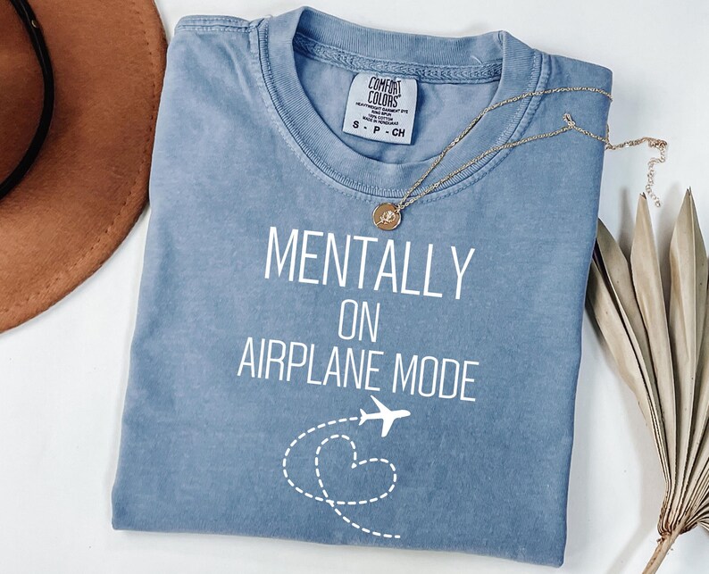 May include: A pale blue t-shirt with the text "MENTALLY ON AIRPLANE MODE" in white. A small airplane graphic and a dotted heart shape are below the text. A gold necklace with a pendant rests on the shirt.
