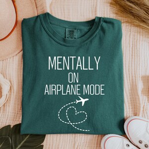 May include: A teal t-shirt with the words "MENTALLY ON AIRPLANE MODE" in white. A white airplane graphic with a dotted heart trail is below the text. The shirt is folded on a cream-coloured surface.