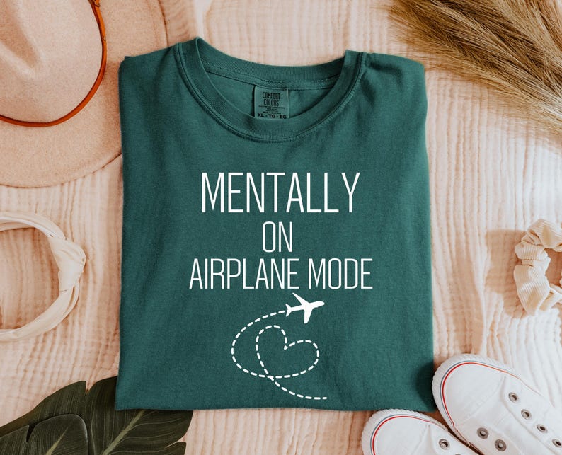 May include: A teal t-shirt with the words "MENTALLY ON AIRPLANE MODE" in white. A white airplane graphic with a dotted heart trail is below the text. The shirt is folded on a cream-coloured surface.
