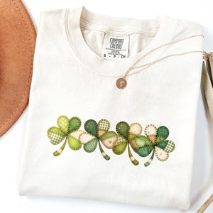 May include: A cream-coloured t-shirt with a stitched shamrock design in shades of green and cream. The shamrocks have a patchwork appearance with various patterns. A gold necklace with a pendant is draped across the shirt.