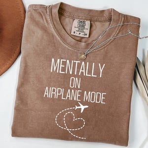 May include: A brown t-shirt with the text "MENTALLY ON AIRPLANE MODE" in white. The design includes a white airplane and a dotted heart shape. A silver necklace with a pendant is on the shirt.
