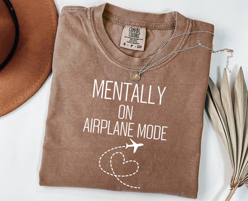 May include: A brown t-shirt with the text "MENTALLY ON AIRPLANE MODE" in white. The design includes a white airplane and a dotted heart shape. A silver necklace with a pendant is on the shirt.