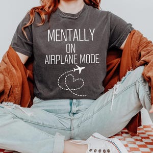May include: Dark grey t-shirt with the text "MENTALLY ON AIRPLANE MODE" in white. A white airplane graphic with a dotted heart trail is below the text. The person is wearing light blue jeans and a brown jacket.