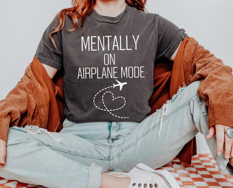 May include: Dark grey t-shirt with the text "MENTALLY ON AIRPLANE MODE" in white. A white airplane graphic with a dotted heart trail is below the text. The person is wearing light blue jeans and a brown jacket.