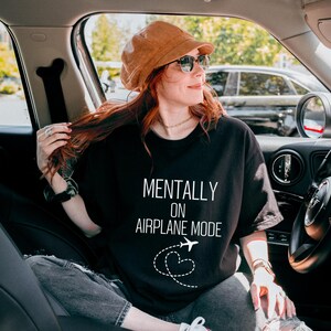 May include: Black t-shirt with the text "MENTALLY ON AIRPLANE MODE" in white, featuring an airplane and heart graphic. The person in the image is wearing a brown corduroy hat and sunglasses, seated in a car.