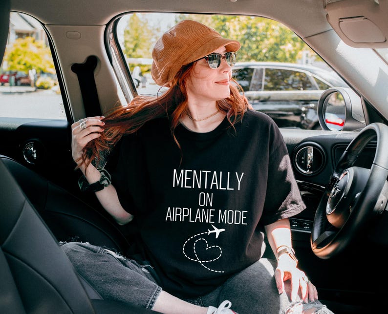 May include: Black t-shirt with the text "MENTALLY ON AIRPLANE MODE" in white, featuring an airplane and heart graphic. The person in the image is wearing a brown corduroy hat and sunglasses, seated in a car.