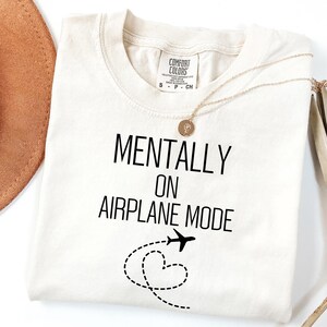 May include: Cream-coloured t-shirt with the text "MENTALLY ON AIRPLANE MODE" in black. A small aeroplane graphic and a dotted heart shape are below the text. A gold necklace and a brown hat are also visible.