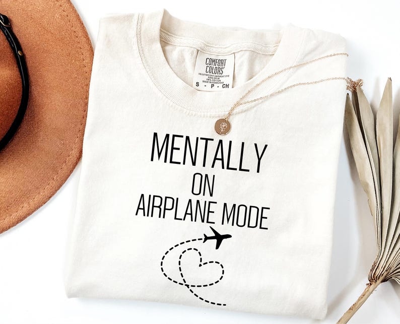 May include: Cream-coloured t-shirt with the text "MENTALLY ON AIRPLANE MODE" in black. A small aeroplane graphic and a dotted heart shape are below the text. A gold necklace and a brown hat are also visible.