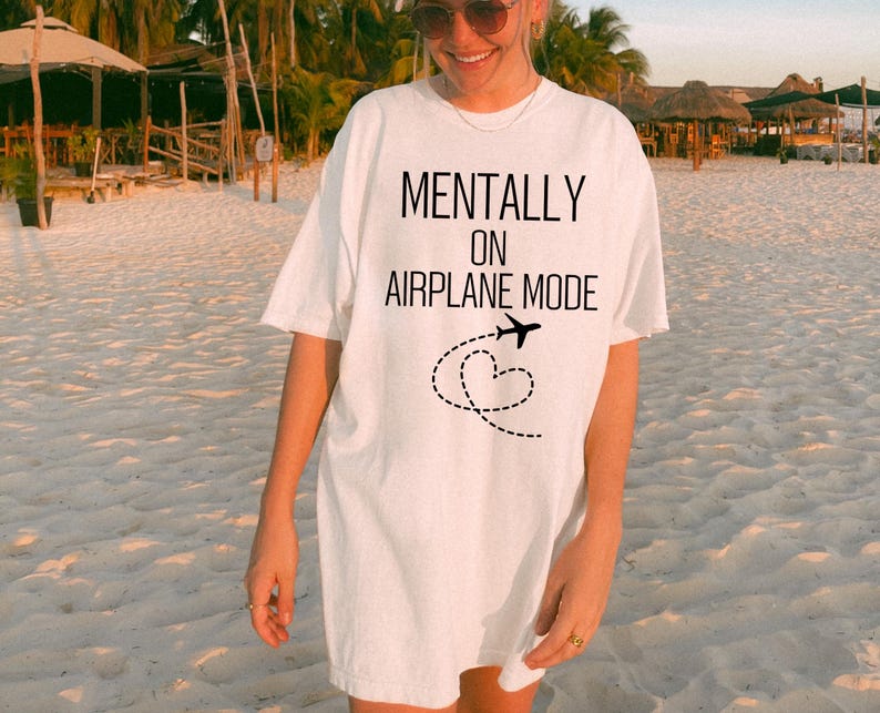 May include: White t-shirt featuring the text "MENTALLY ON AIRPLANE MODE" in black. A black airplane graphic with a dotted heart-shaped trail is below the text. The shirt has a loose fit.