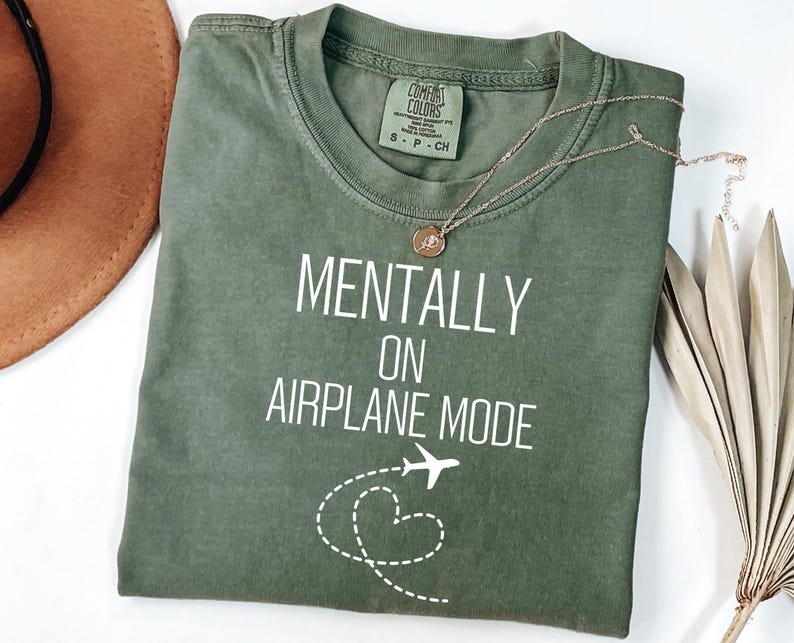 May include: An olive green t-shirt with the text "MENTALLY ON AIRPLANE MODE" in white. A small airplane graphic and a dotted heart design are below the text. A gold necklace with a pendant is on the shirt.