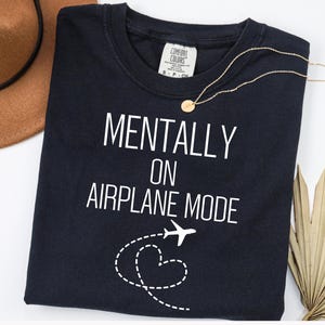 May include: A navy blue t-shirt with the words "MENTALLY ON AIRPLANE MODE" in white. A gold necklace and a brown hat are also in the image. A white airplane graphic with a dotted heart trail is below the text.