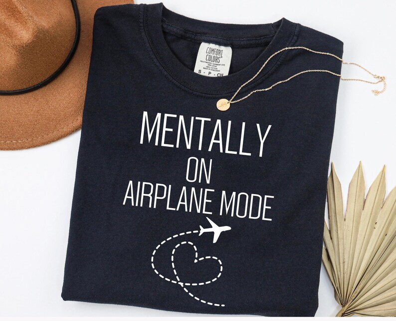 May include: A navy blue t-shirt with the words "MENTALLY ON AIRPLANE MODE" in white. A gold necklace and a brown hat are also in the image. A white airplane graphic with a dotted heart trail is below the text.
