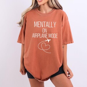 May include: Burnt orange t-shirt with the text "MENTALLY ON AIRPLANE MODE" in white. A white airplane graphic with a dotted heart-shaped trail is below the text. The shirt has short sleeves and a relaxed fit.