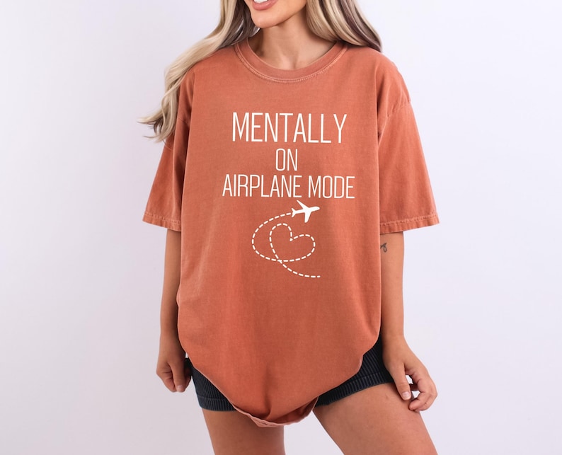 May include: Burnt orange t-shirt with the text "MENTALLY ON AIRPLANE MODE" in white. A white airplane graphic with a dotted heart-shaped trail is below the text. The shirt has short sleeves and a relaxed fit.