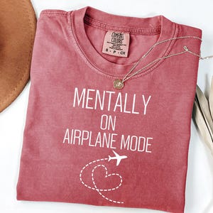 May include: A red-toned t-shirt with the text "MENTALLY ON AIRPLANE MODE" in white. A small airplane graphic and a dotted heart design are below the text. A silver necklace with a pendant is on the shirt.
