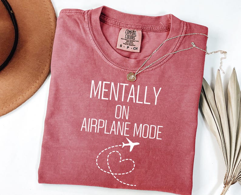 May include: A red-toned t-shirt with the text "MENTALLY ON AIRPLANE MODE" in white. A small airplane graphic and a dotted heart design are below the text. A silver necklace with a pendant is on the shirt.