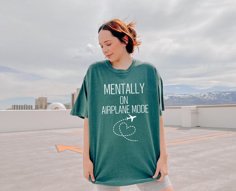 May include: A teal green t-shirt with the words "MENTALLY ON AIRPLANE MODE" in white, and a white airplane and heart graphic. The shirt has a relaxed fit and is made of a soft material.