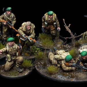 May include: Miniature figurines of soldiers in tan uniforms and green berets, posed in a diorama setting. The soldiers are armed with rifles and machine guns, some kneeling or prone among the grass and foliage. The scene is set on a dark, round base.
