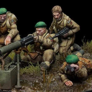 May include: Miniature figures of soldiers in tan uniforms and green berets, operating a machine gun. One soldier kneels, aiming the weapon, while others observe or prepare. The scene is set on a small, detailed diorama.