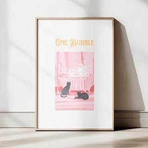 Pode incluir: Impressão de arte emoldurada com uma ilustração rosa e branca. A obra retrata uma banheira cheia de bolhas e dois gatos pretos, com o texto "ONE BUBBLE". A moldura é de cor de madeira clara.